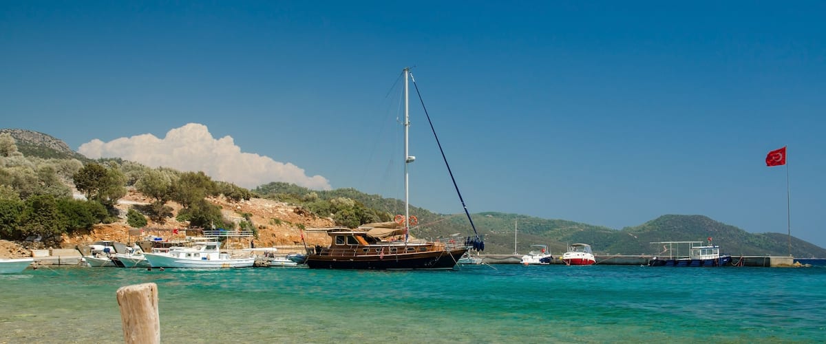 A coastal scene with boats docked near a rocky shore, a flag, and hills in the background.