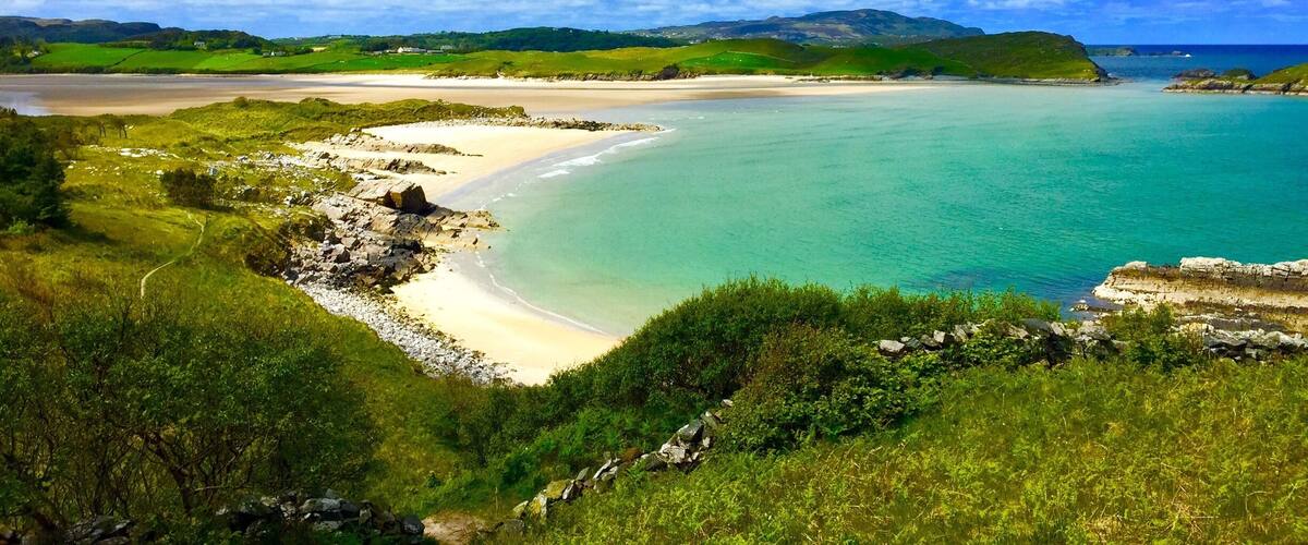 A coastal landscape with a sandy beach, turquoise waters, and green hills under a partly cloudy sky.