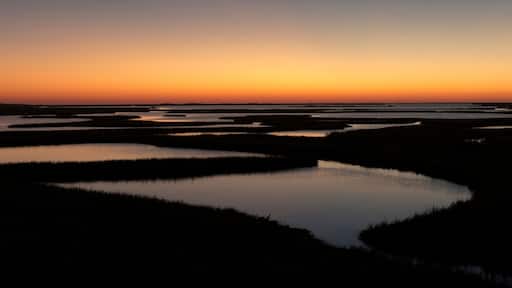 A sunset over a marshy landscape with water bodies and distant hills.
