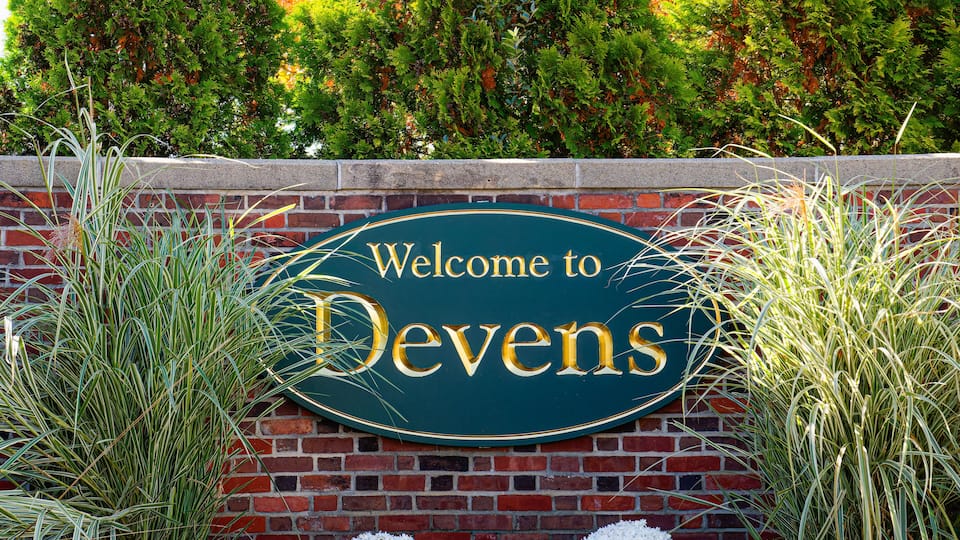 A welcome sign to Devens with a brick wall and greenery in the background.