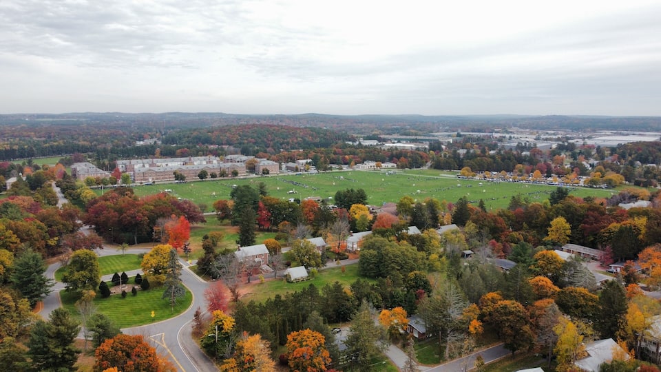 An aerial view of a campus with a large green field, surrounded by autumn-colored trees and residential buildings.