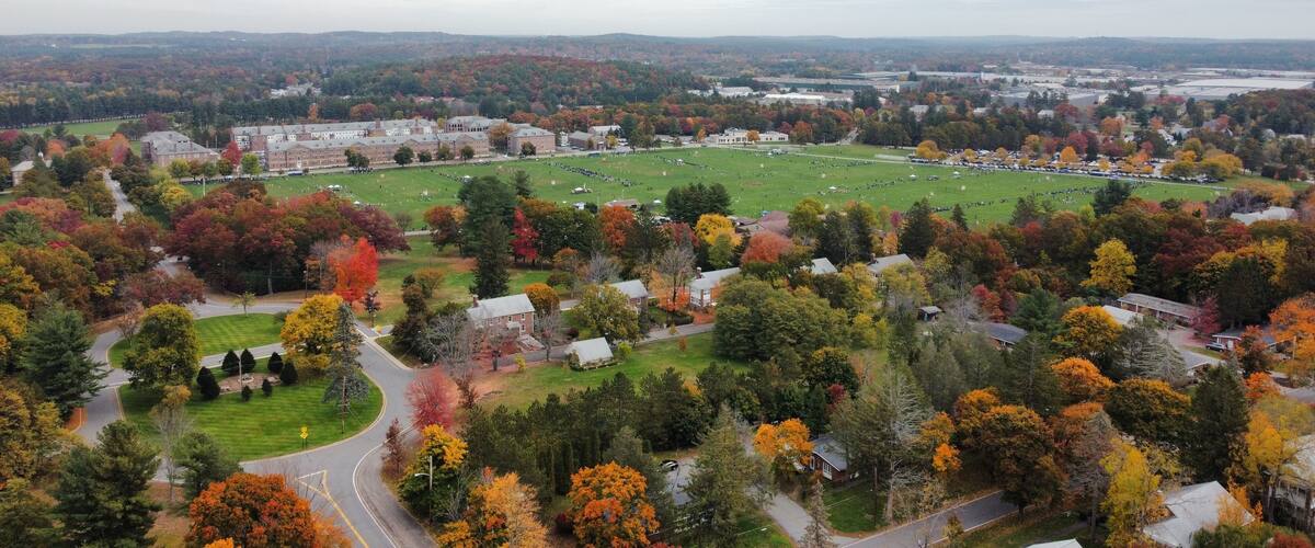 An aerial view of a campus with a large green field, surrounded by autumn-colored trees and residential buildings.
