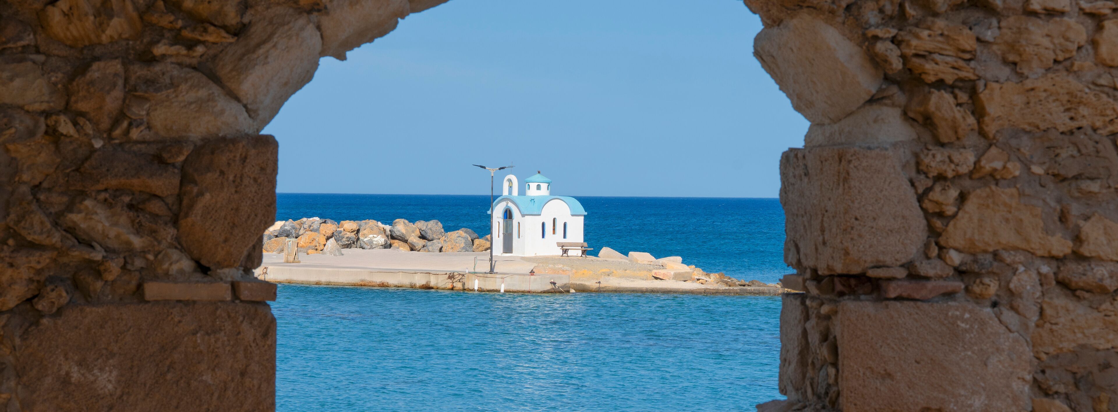 A coastal view through stone arches, featuring a small white church with a blue dome and a bench on a rocky shore.