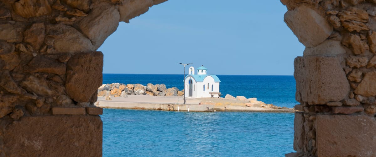 A coastal view through stone arches, featuring a small white church with a blue dome and a bench on a rocky shore.