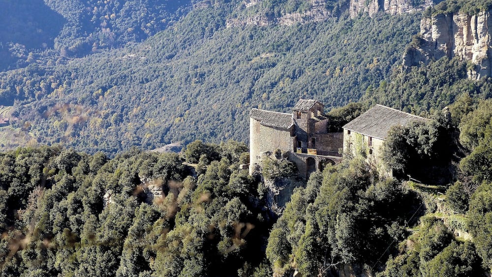 A medieval stone castle perched on a hillside, surrounded by dense forest and rugged mountains.