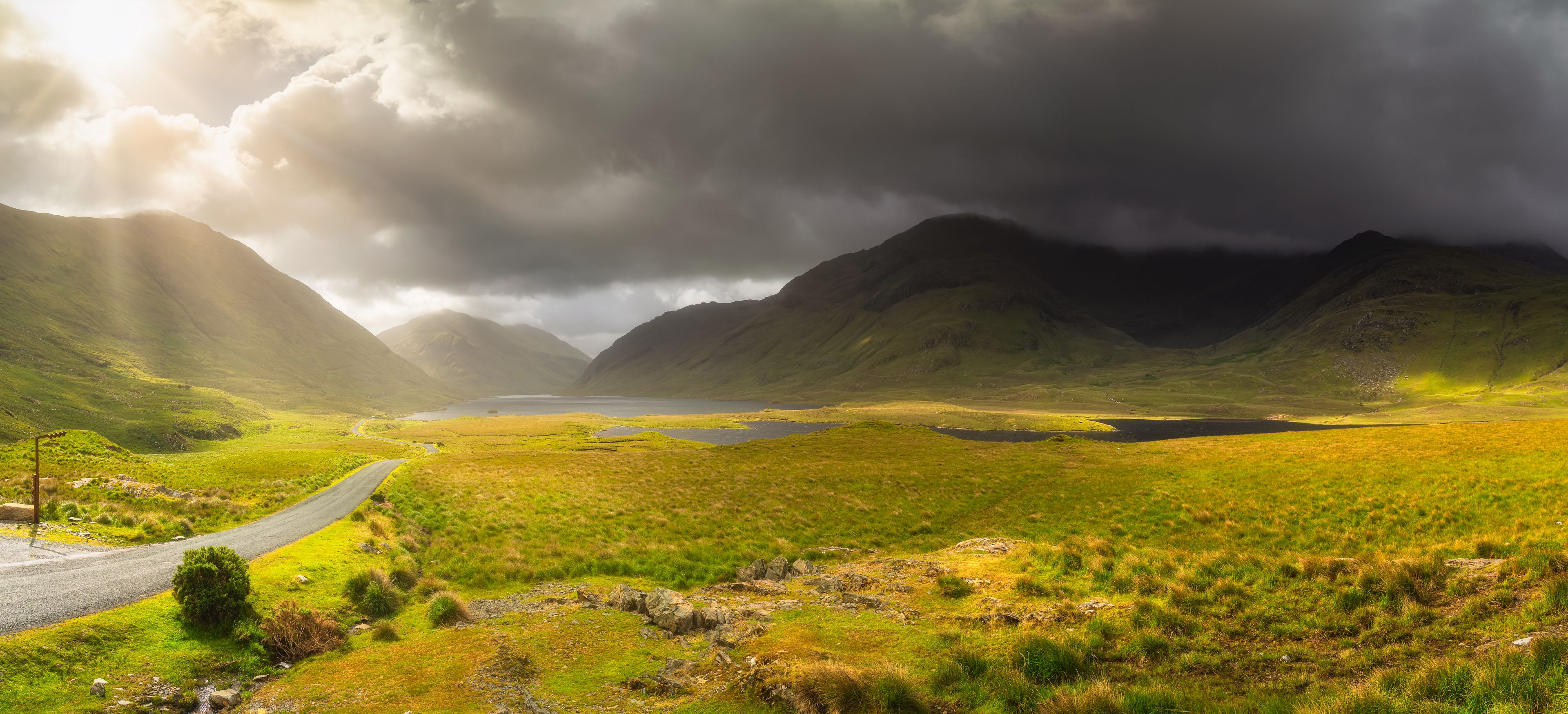 Panorama of Doolough Valley with lakes, Glenummera and Glencullin mountain ranges illuminated by sunlight, dark dramatic sky, County Mayo, Ireland