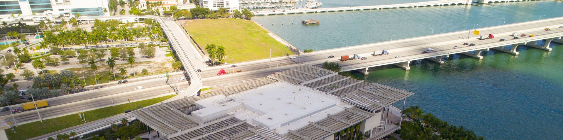 Vista aérea de una zona urbana costera con un edificio grande, palmeras, un puente y un puerto deportivo.