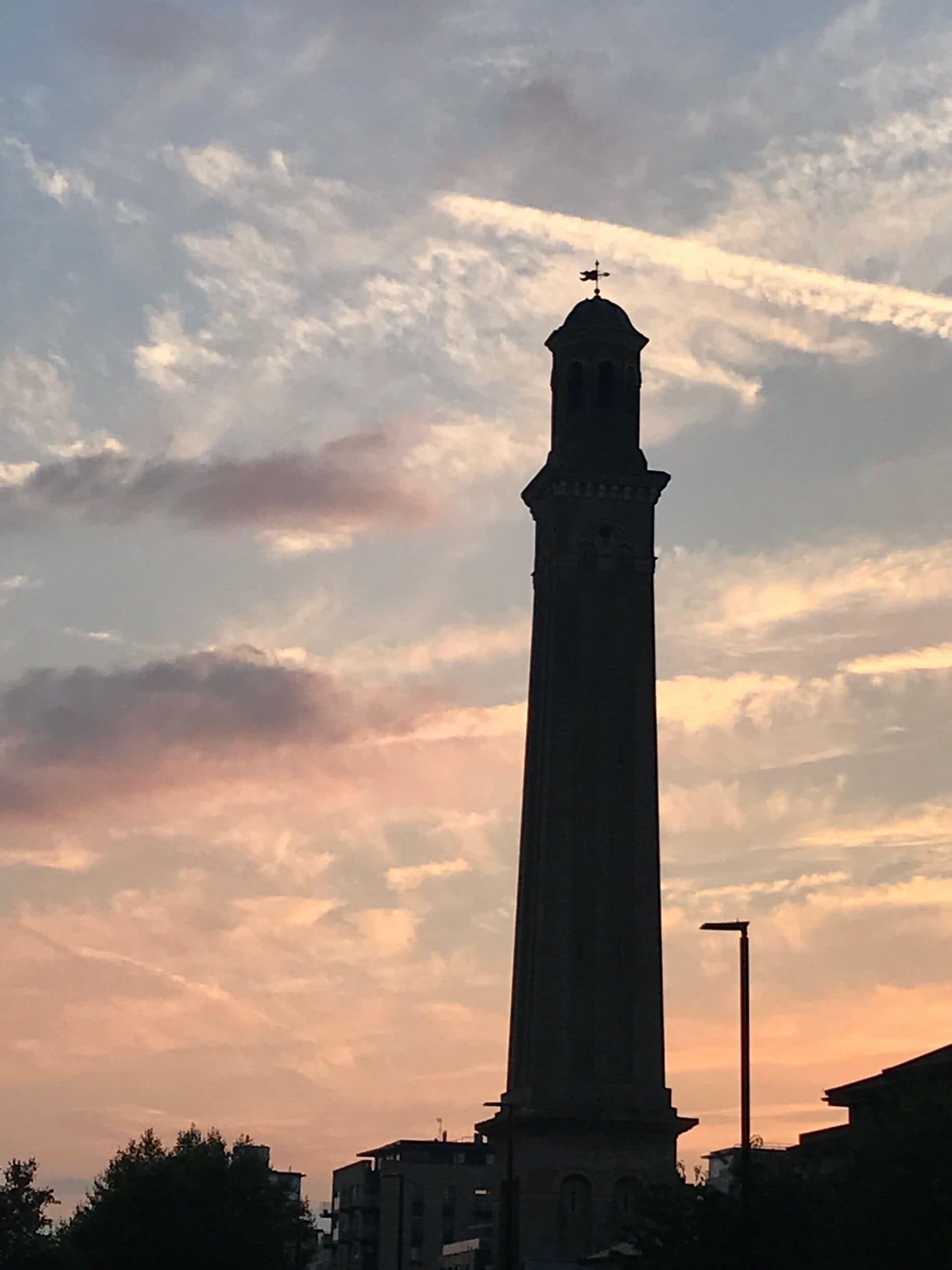 Korkea torni, jonka huipulla on tuulikukko, ja taustalla auringonlaskun värittämä taivas pilvineen.