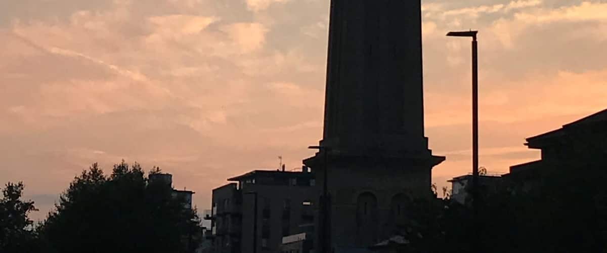 Korkea torni, jonka huipulla on tuulikukko, ja taustalla auringonlaskun värittämä taivas pilvineen.