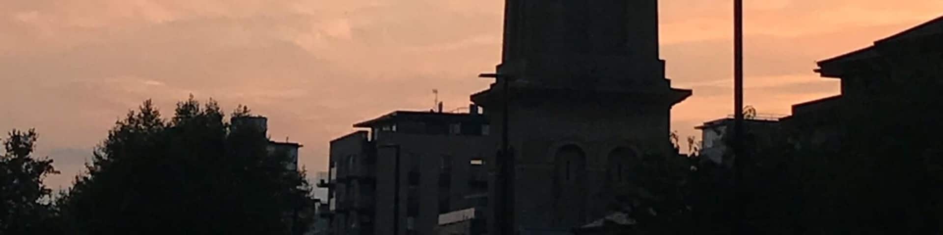 A tall tower with a weather vane at the top, set against a sunset sky with clouds.