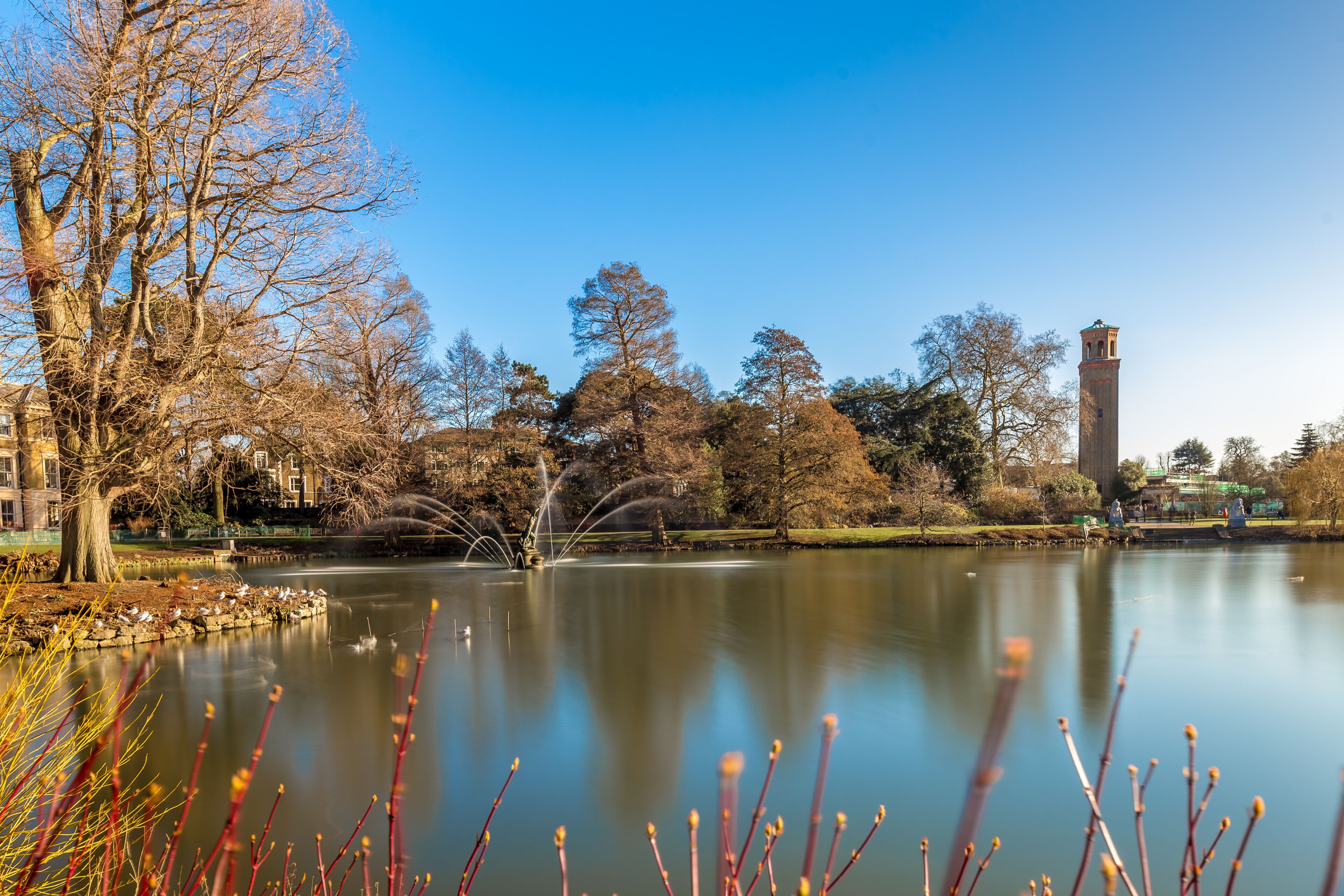 Um lago tranquilo com uma fonte, rodeado por árvores e uma torre histórica.
