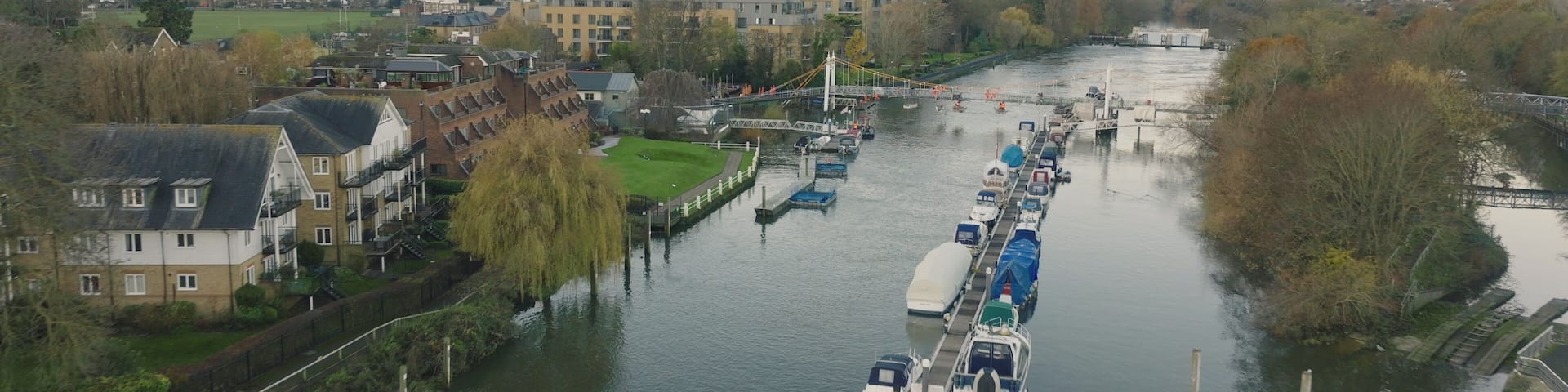 London, Teddington: Boats moored along the River Thames in Teddington, London city, create a serene scene with residential buildings houses and trees under a cloudy sky. Drone flight