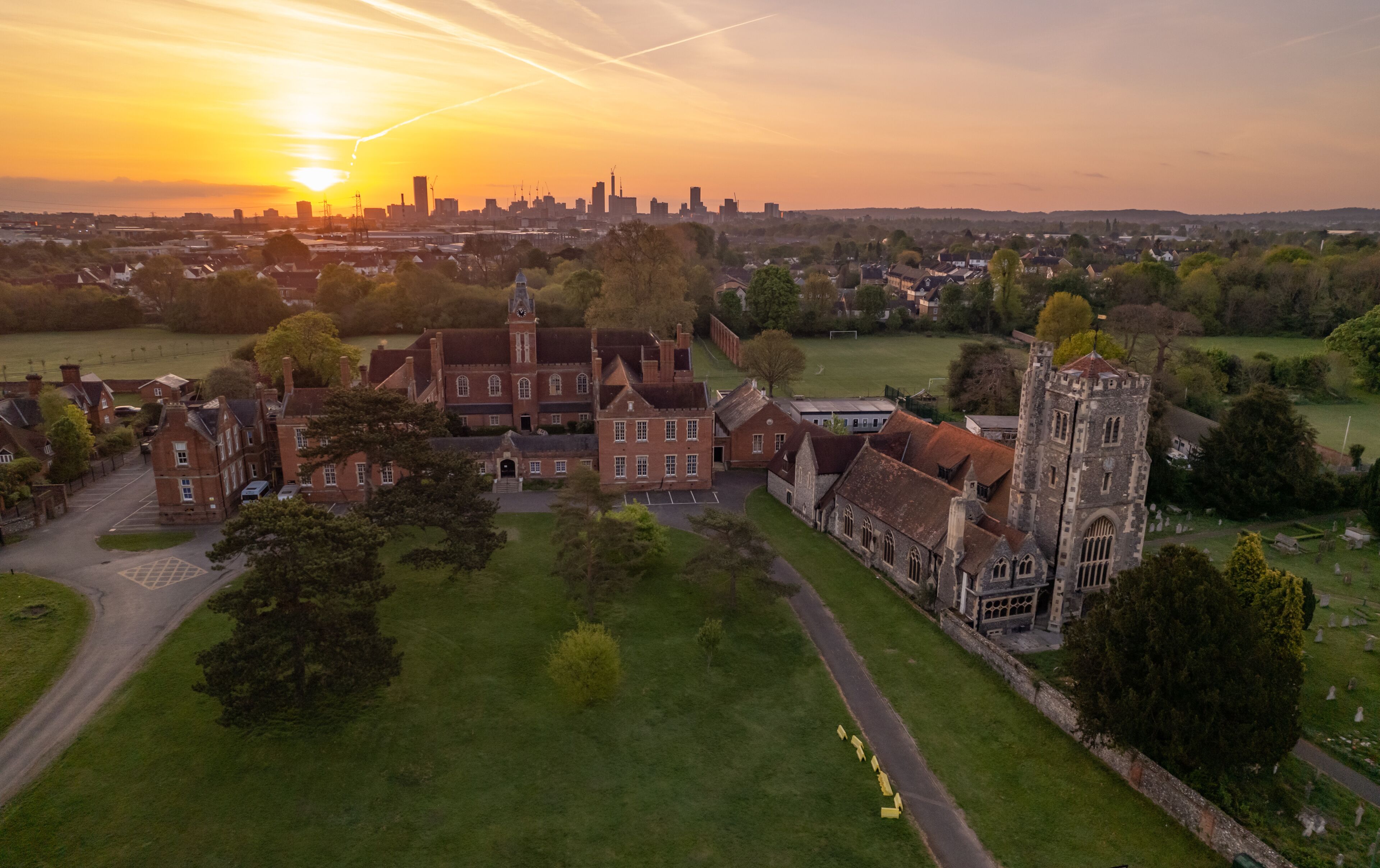 A historic building complex with a prominent tower, surrounded by green fields and a city skyline in the background.