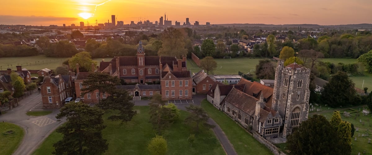 A historic building complex with a prominent tower, surrounded by green fields and a city skyline in the background.