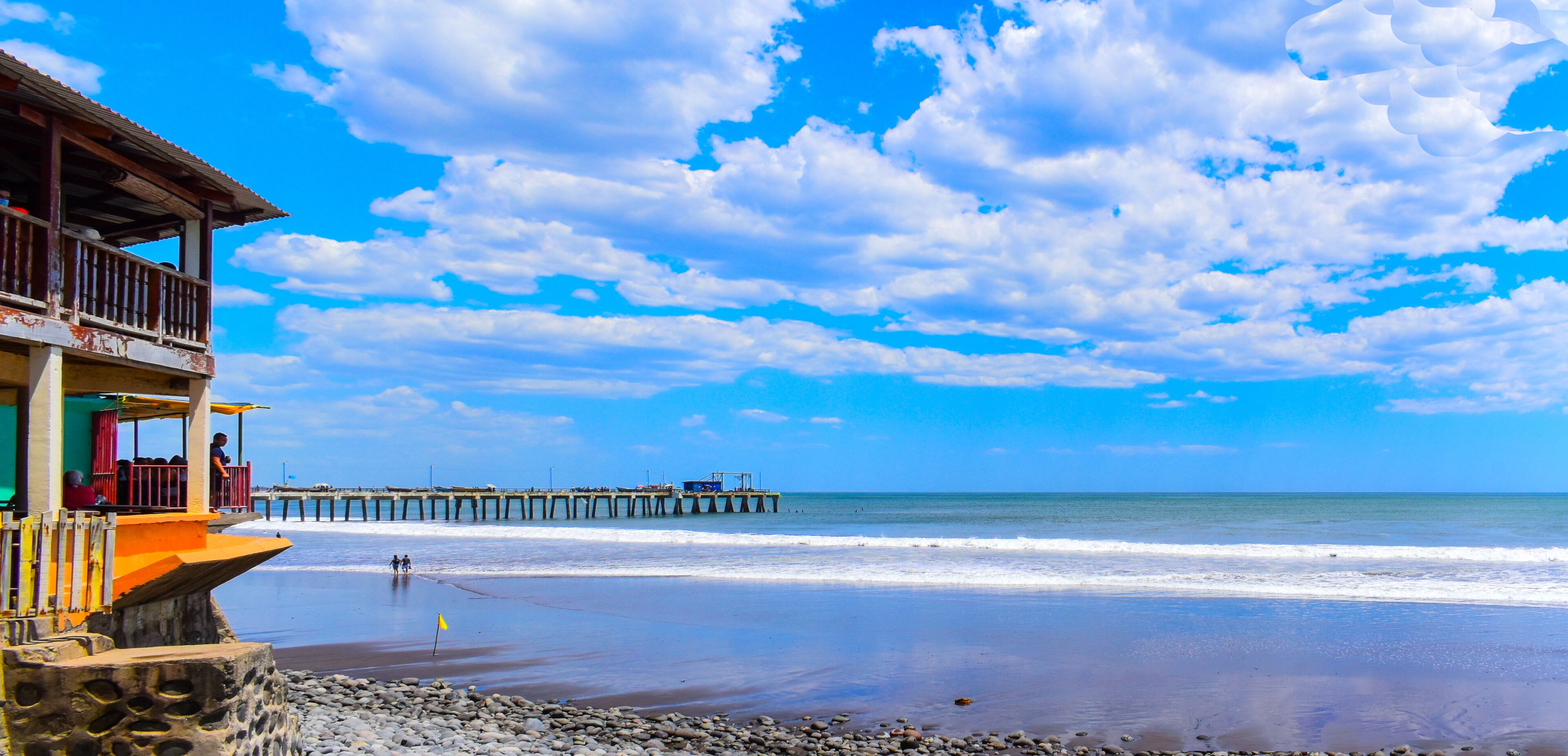 A beach with a wooden pier extending into the ocean, a rocky shoreline, and a clear blue sky with scattered clouds.