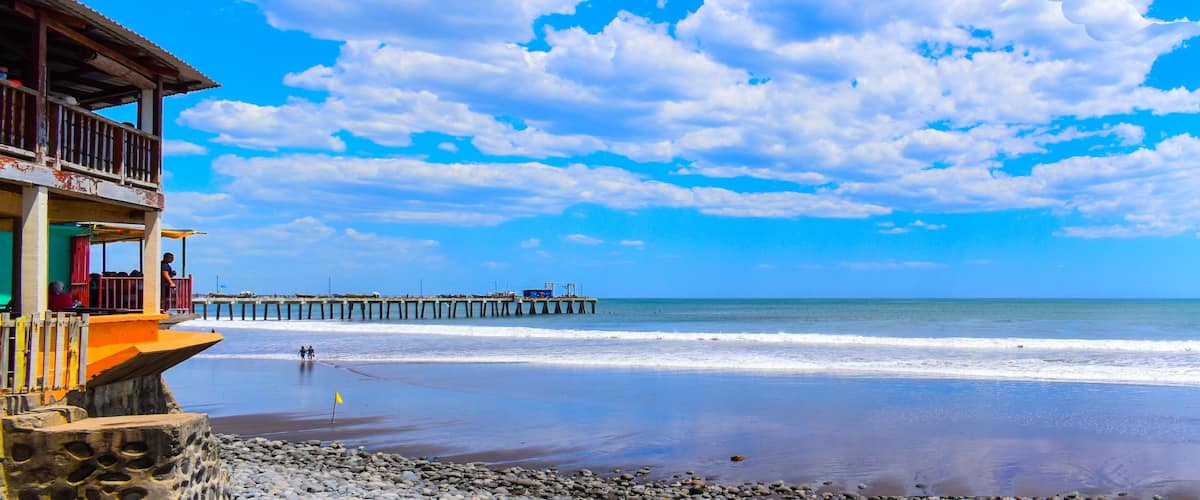 A beach with a wooden pier extending into the ocean, a rocky shoreline, and a clear blue sky with scattered clouds.