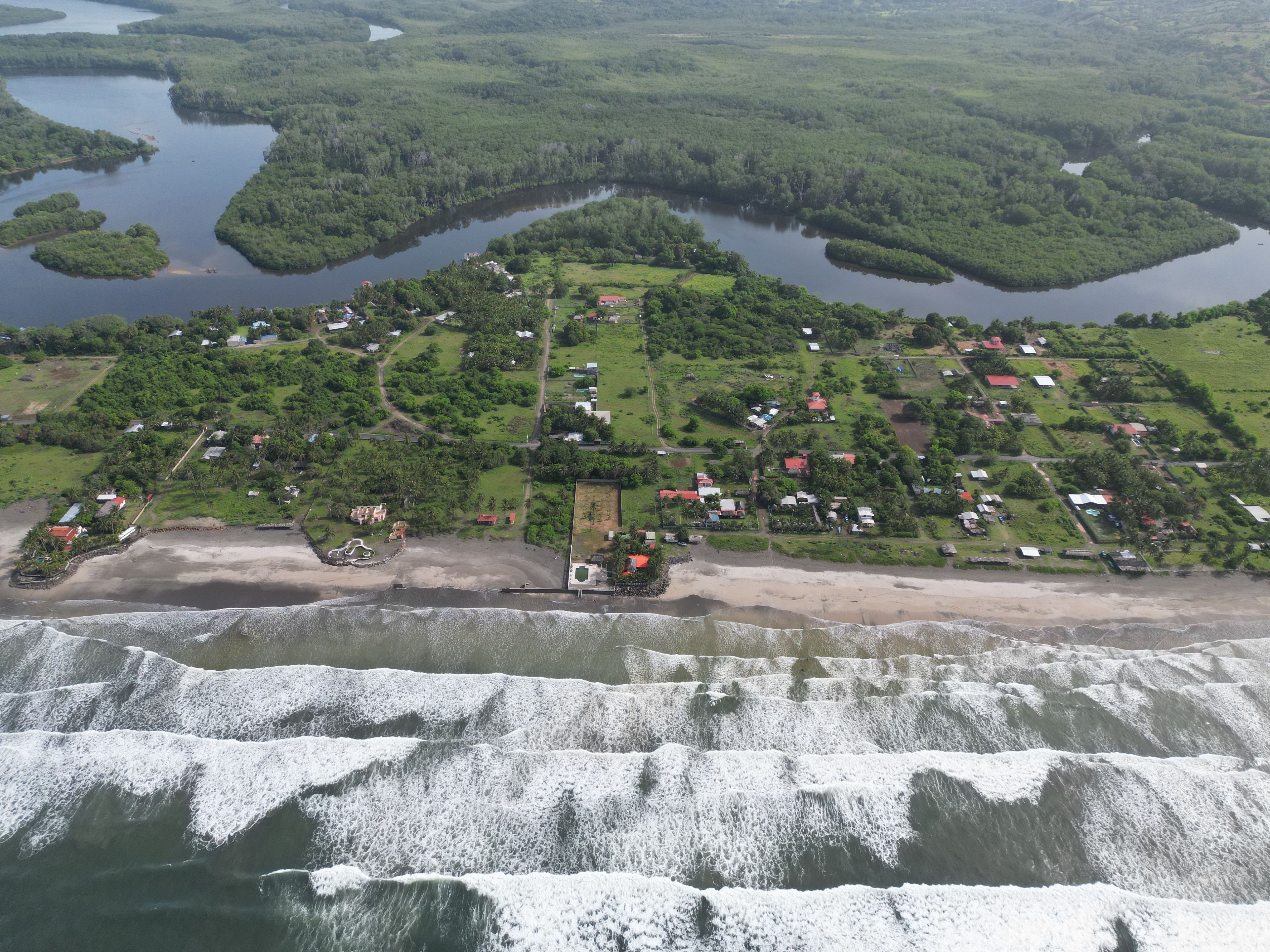Vista aérea de una zona costera con playa, olas y un asentamiento denso con casas y vegetación.