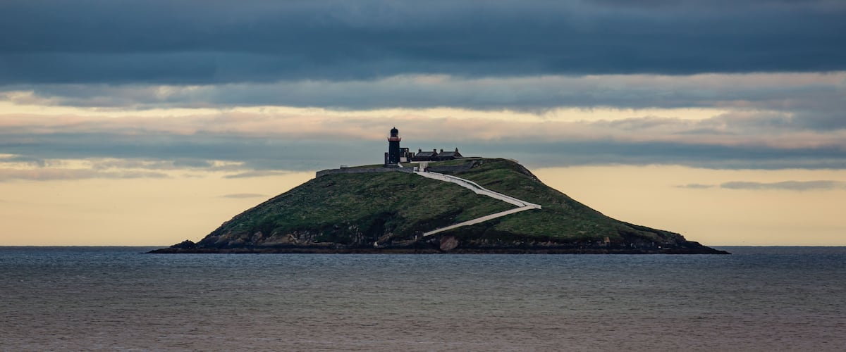 A lighthouse on a grassy hill by the sea under a cloudy sky.
