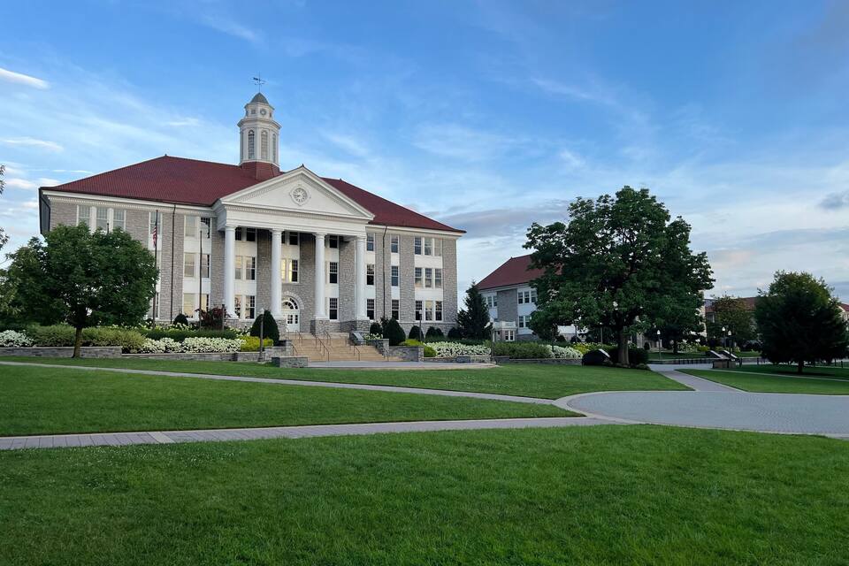 A large, historic building with a red roof and columns, surrounded by well-maintained lawns and trees.