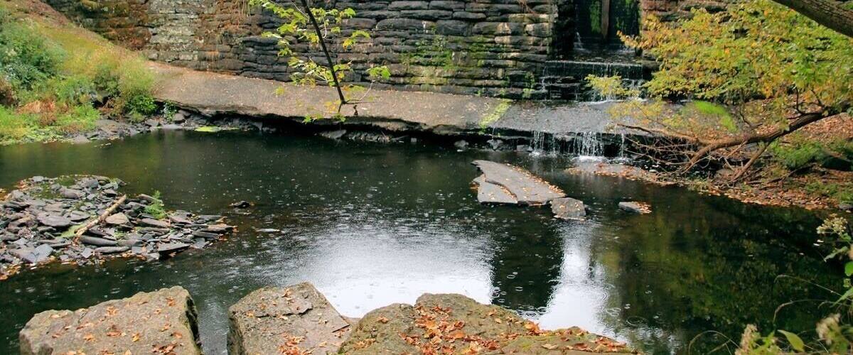 A stone bridge over a calm river, surrounded by autumn foliage and a small waterfall.
