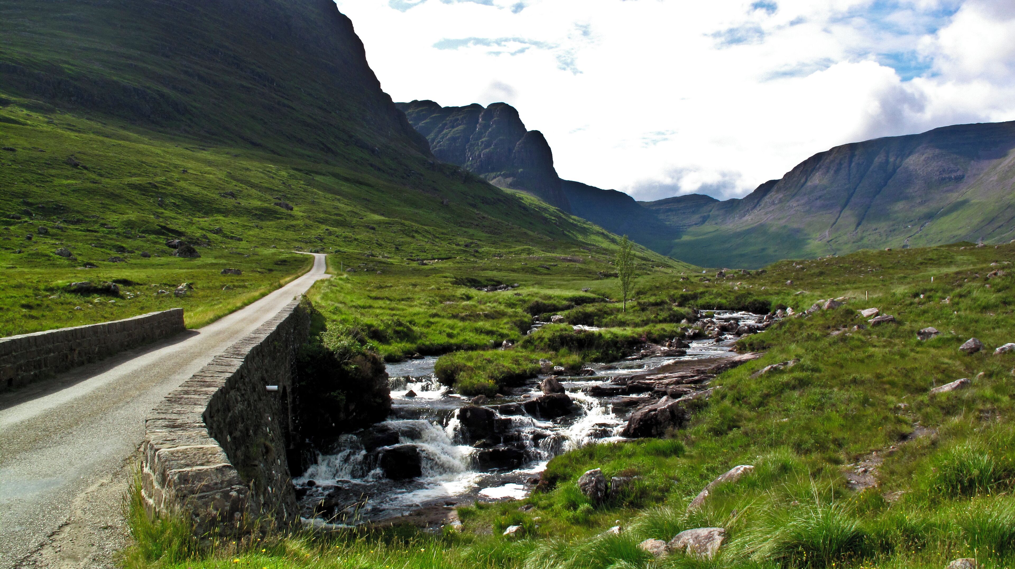 A winding road through a mountainous landscape with a stream running alongside.