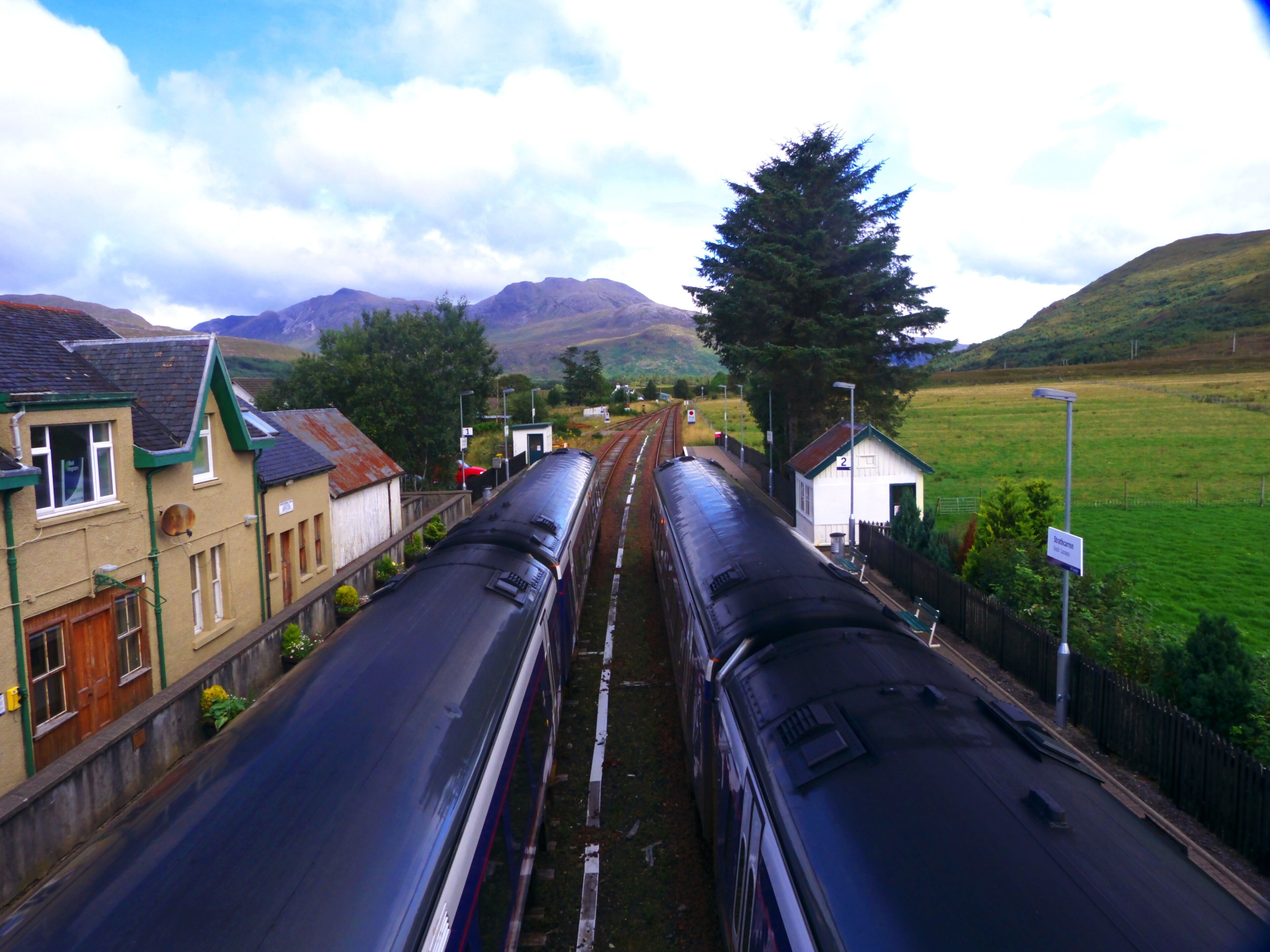 A railway station with two trains on adjacent tracks, surrounded by houses and a green field with mountains in the background.
