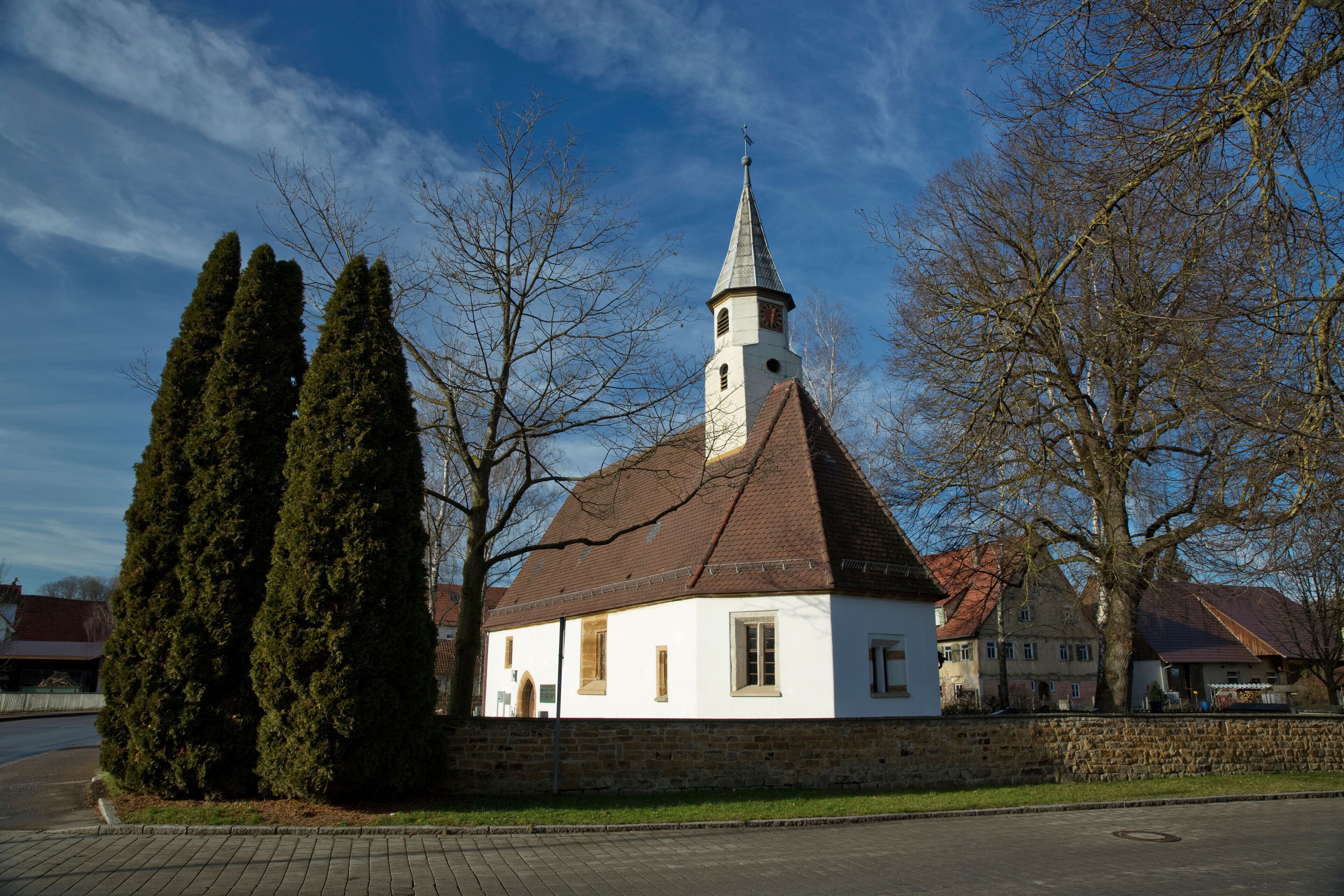 A white church with a pointed steeple and a clock tower, surrounded by trees and a stone wall.