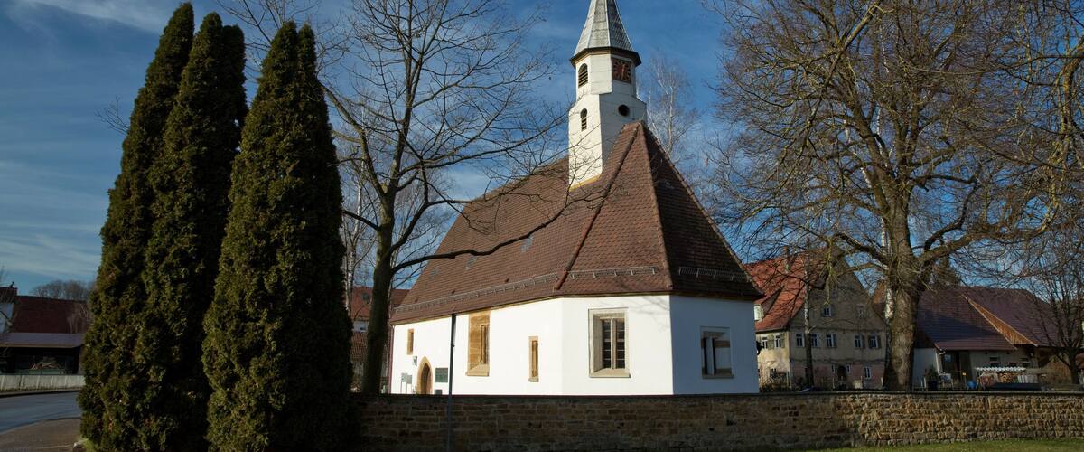 A white church with a pointed steeple and a clock tower, surrounded by trees and a stone wall.