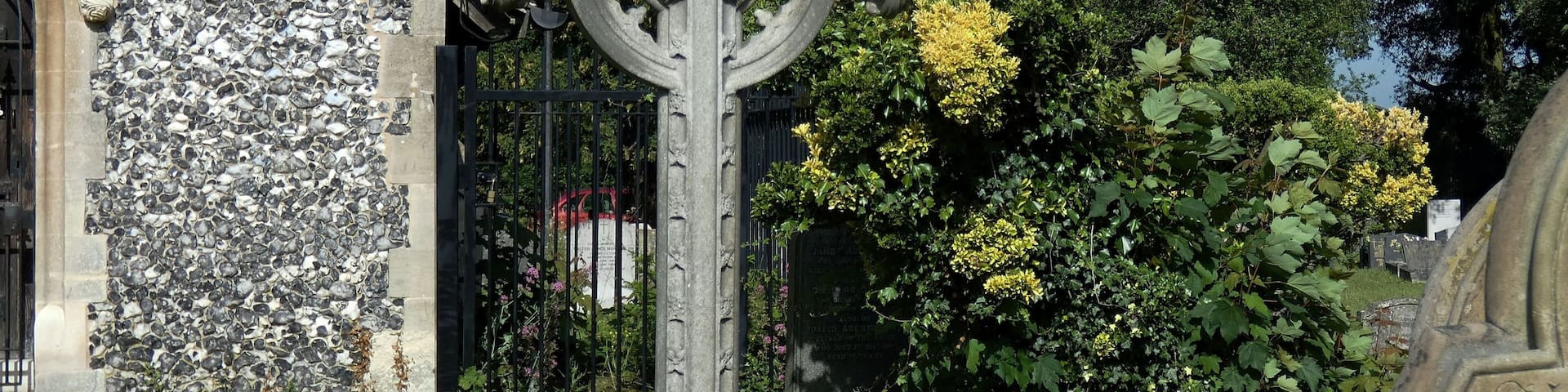A stone cross with a circular design, surrounded by greenery and a stone wall with a patterned surface.