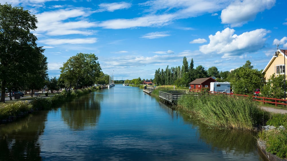 A scenic view of Göta Canal with calm waters reflecting a bright blue sky and white clouds. Lush greenery, traditional Swedish houses, and a peaceful summer atmosphere. Töreboda, Sweden