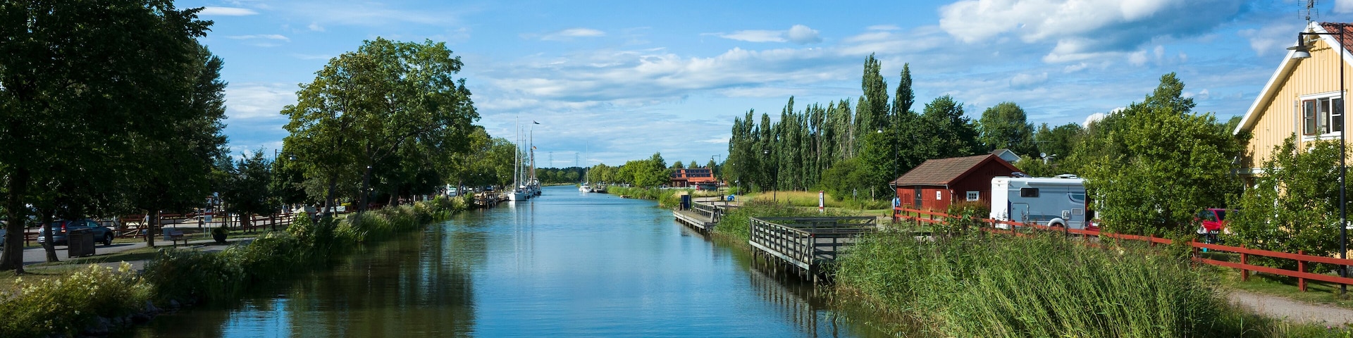 A scenic view of Göta Canal with calm waters reflecting a bright blue sky and white clouds. Lush greenery, traditional Swedish houses, and a peaceful summer atmosphere. Töreboda, Sweden