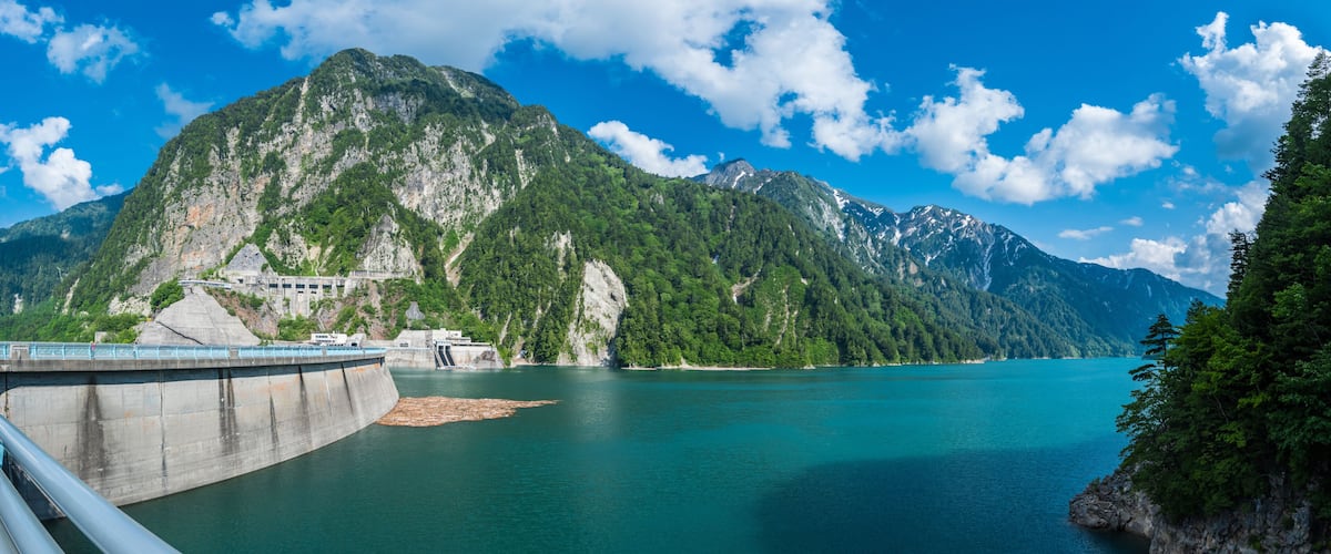 A dam with a bridge, a calm lake, and a mountainous backdrop.