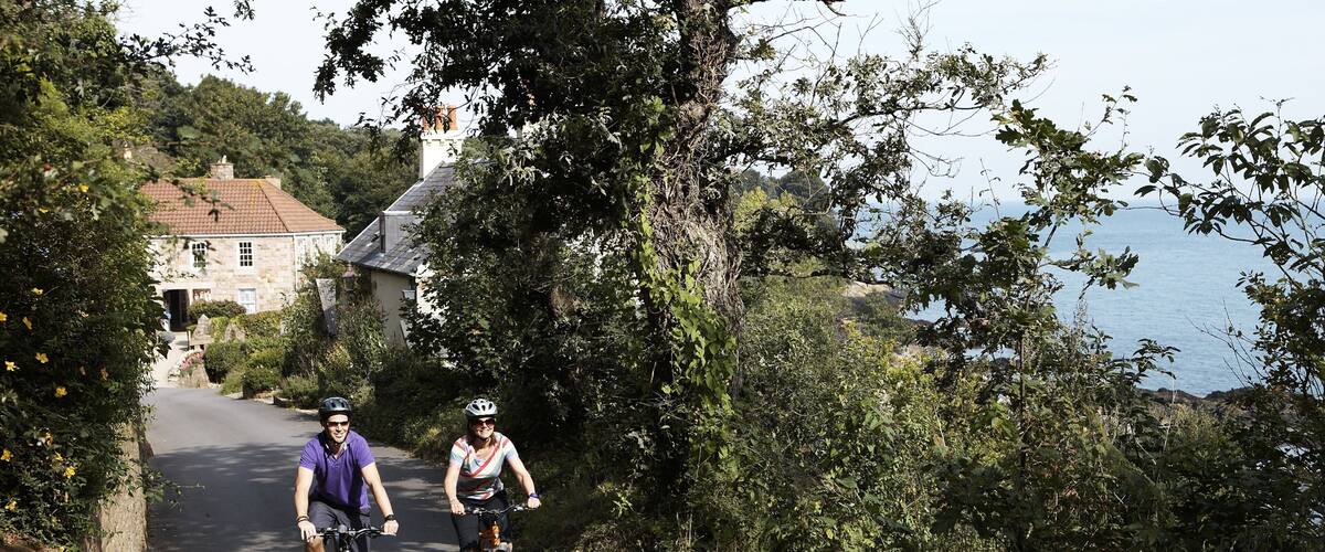 Two cyclists on a narrow road flanked by trees and a stone building.