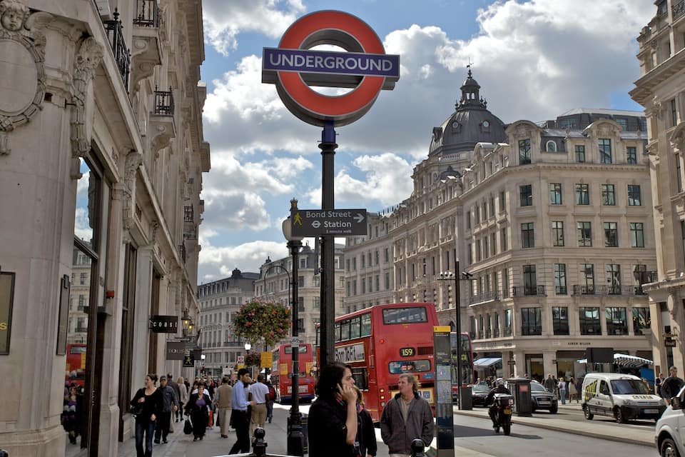 Eine belebte Straße mit einem auffälligen Hinweisschild der London Underground, historischen Gebäuden und einem roten Doppeldeckerbus.