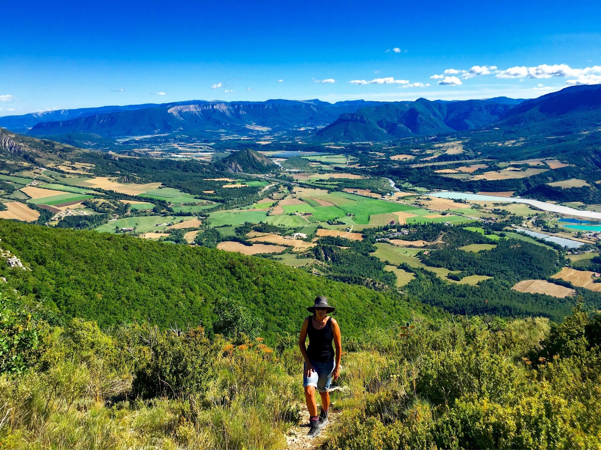 A hiker on a trail overlooking a valley with fields, mountains, and a river.