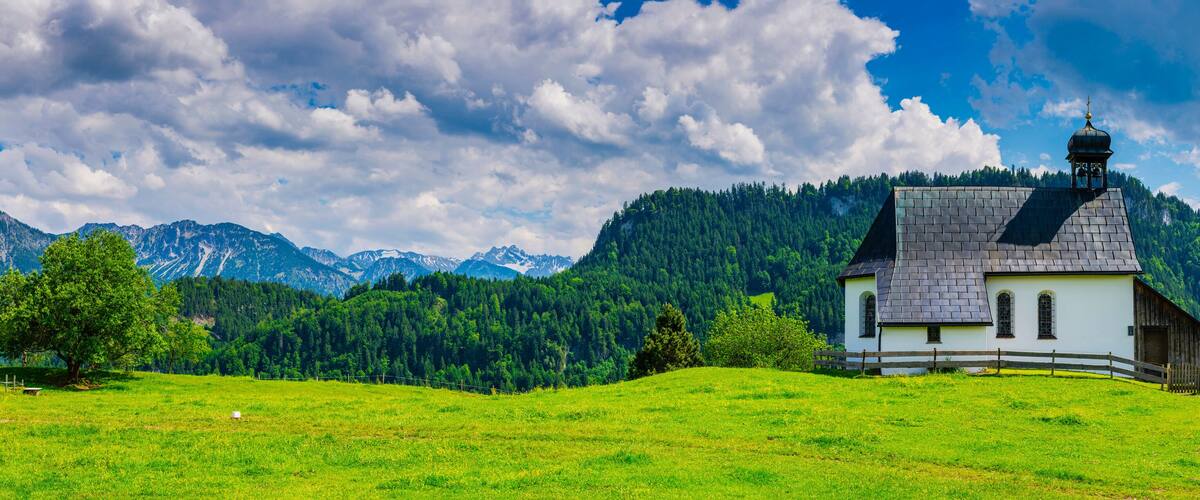A small white church with a dark roof and a bell tower, surrounded by lush green fields and mountains in the background.