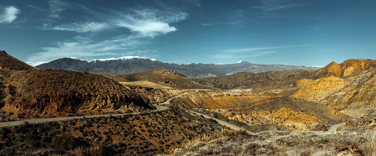 A mountainous landscape with a winding road, rocky terrain, and distant snow-capped peaks.