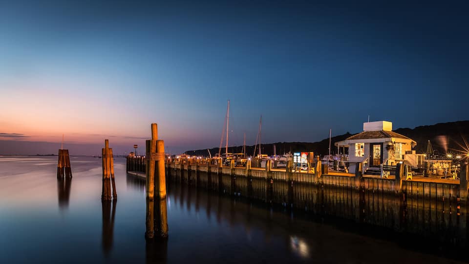 A waterfront at dusk with wooden pilings, a dock, and a small building with lights.