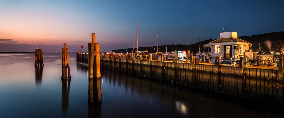 A waterfront at dusk with wooden pilings, a dock, and a small building with lights.