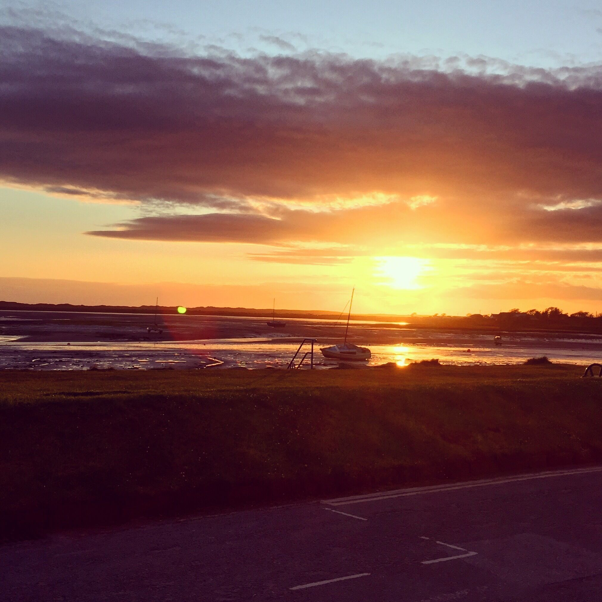A sunset over a coastal area with boats on the water and a road in the foreground.