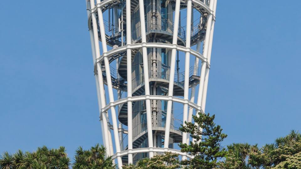 A tall, cylindrical tower with multiple balconies and a glass facade, surrounded by greenery.