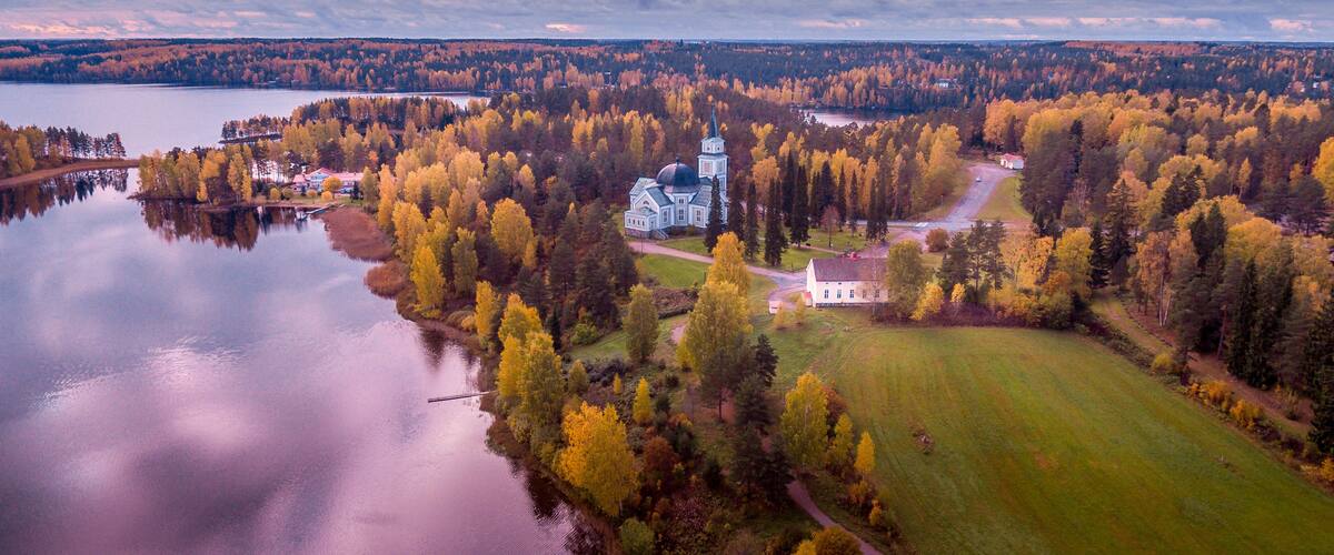 An aerial view of a lakeside estate with a prominent church, surrounded by autumn-colored trees and a calm lake.