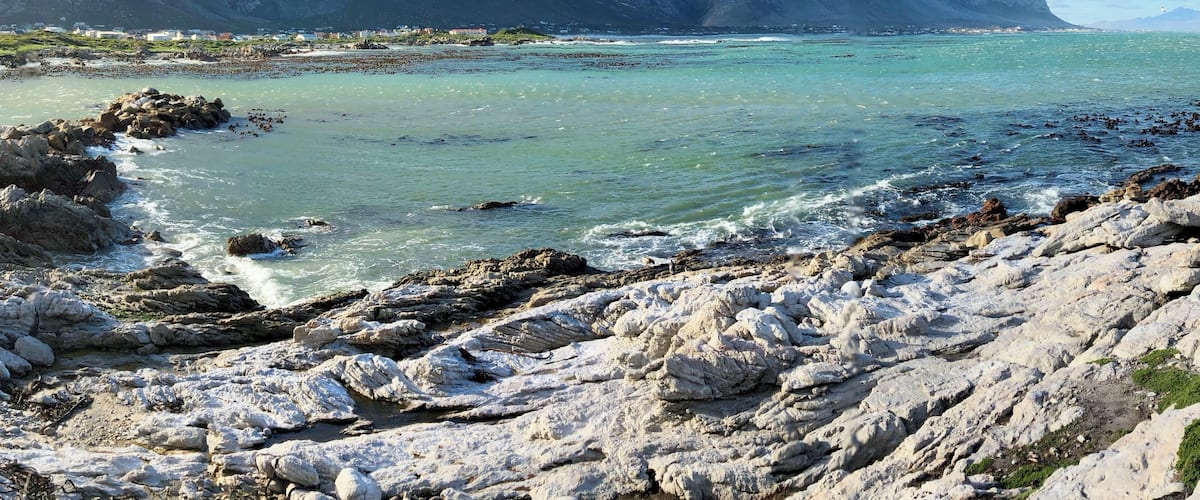 A coastal landscape with rocky shores, clear turquoise waters, and distant mountains under a partly cloudy sky.