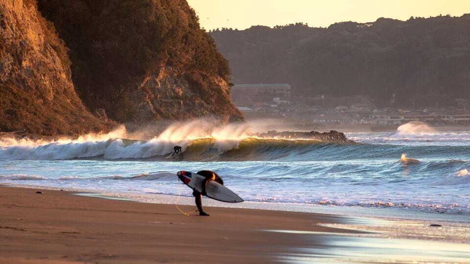 A surfer walking on a beach with waves crashing against a rocky cliff.