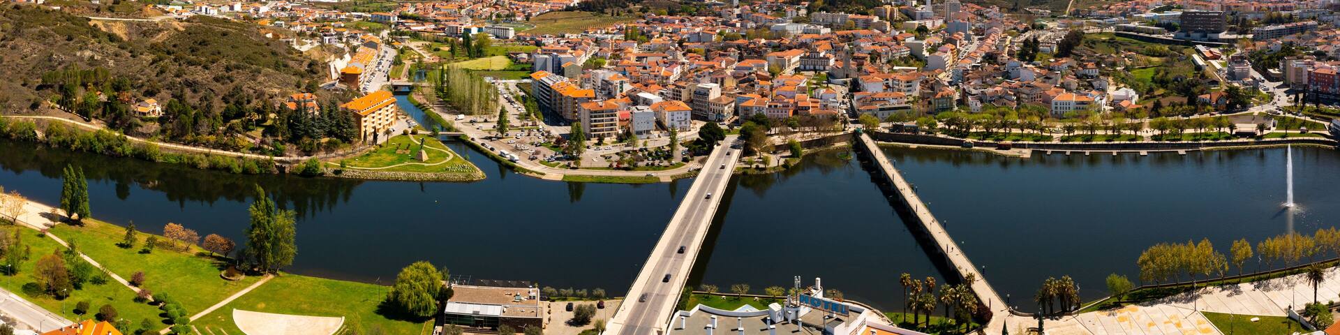 Scenic panorama of Mirandela cityscape on banks of Tua overlooking residential districts, two bridges across river and gentle mountain slopes covered with forests in background on spring day, Portugal