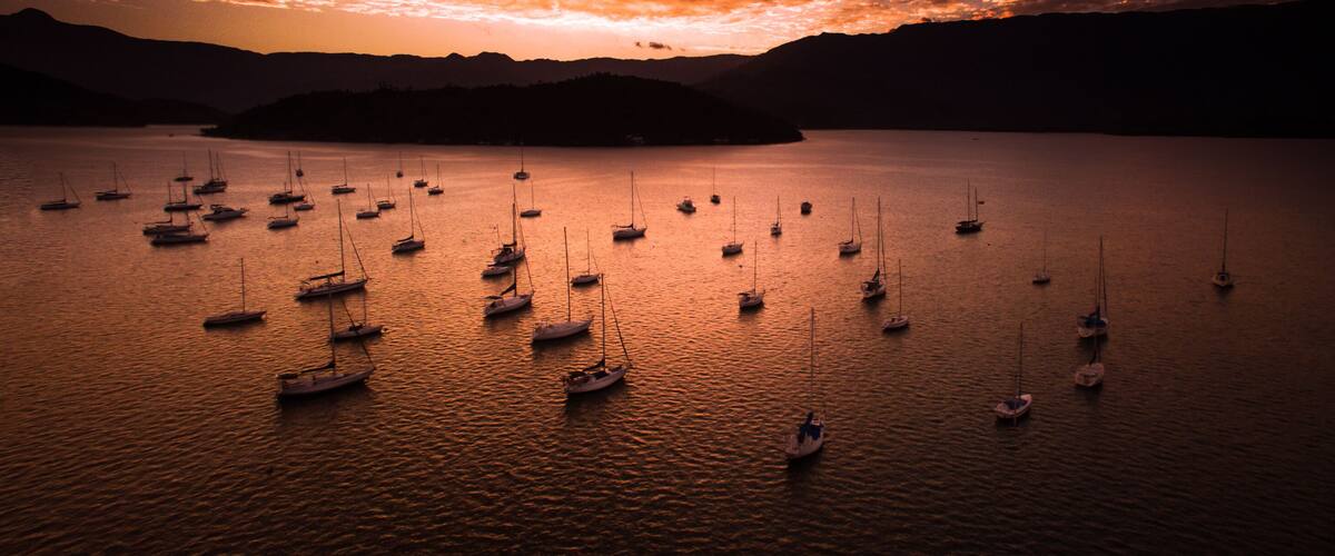 A calm lake with numerous sailboats anchored, surrounded by mountains under a dramatic sunset sky.