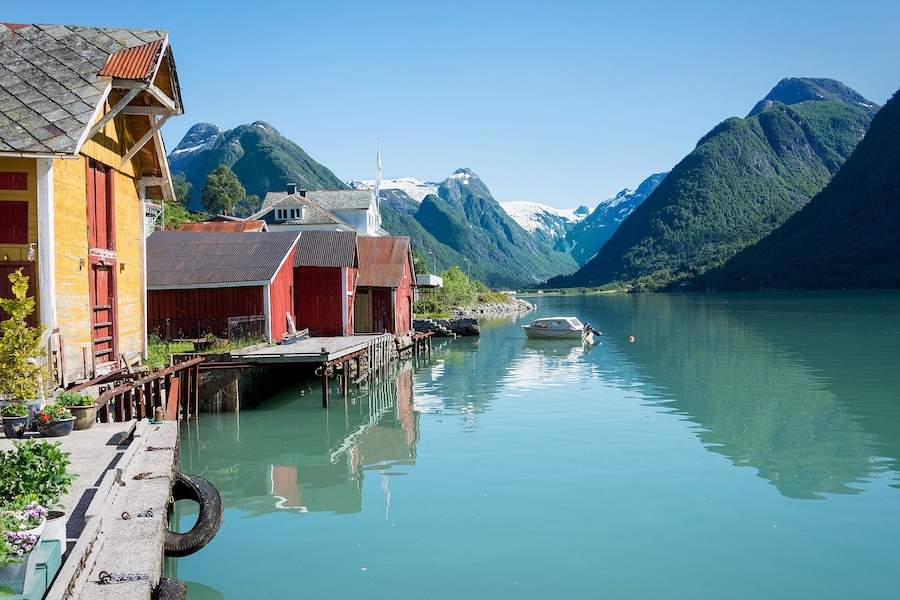 View over the fjord Fjaerlandsfjord and the village of Mundal (or Fjaerland) with some snow-capped mountains, a boat, reflection in the water and yellow and red boathouses in Sogn of Fjordane, Norway.