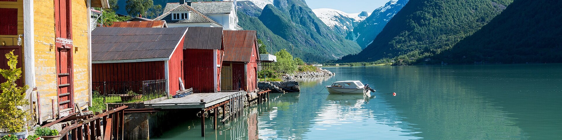 View over the fjord Fjaerlandsfjord and the village of Mundal (or Fjaerland) with some snow-capped mountains, a boat, reflection in the water and yellow and red boathouses in Sogn of Fjordane, Norway.