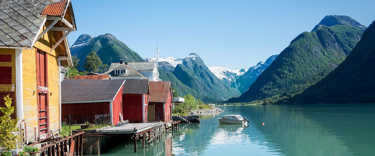 A calm lake with a wooden dock, red buildings, and snow-capped mountains in the background.
