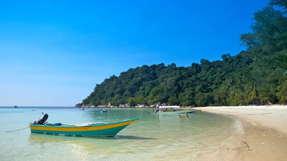A beach with clear turquoise water, a small boat, and a forested hill in the background.